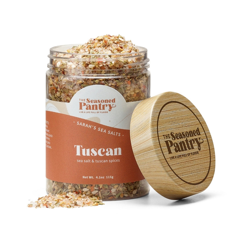 A clear glass jar with a wooden lid containing medium grain seasoning blend, labeled as 'Tuscan Salt' from The Seasoned Pantry.